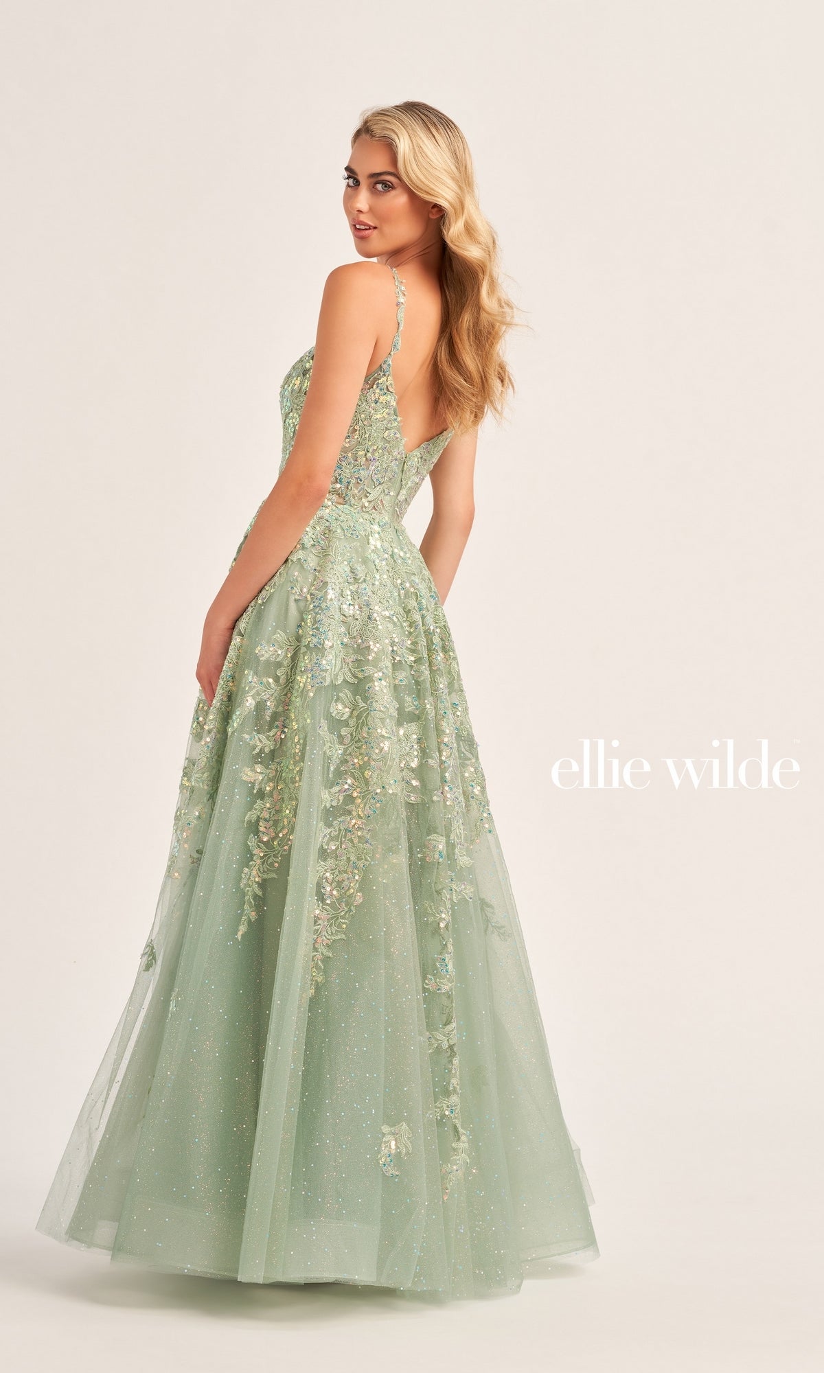 A woman in a pale green Glitter-Tulle Ellie Wilde Long Prom Dress EW35114 with a fitted bodice and thin straps stands slightly turned, highlighting the embellished back and elegant floral appliqué details.